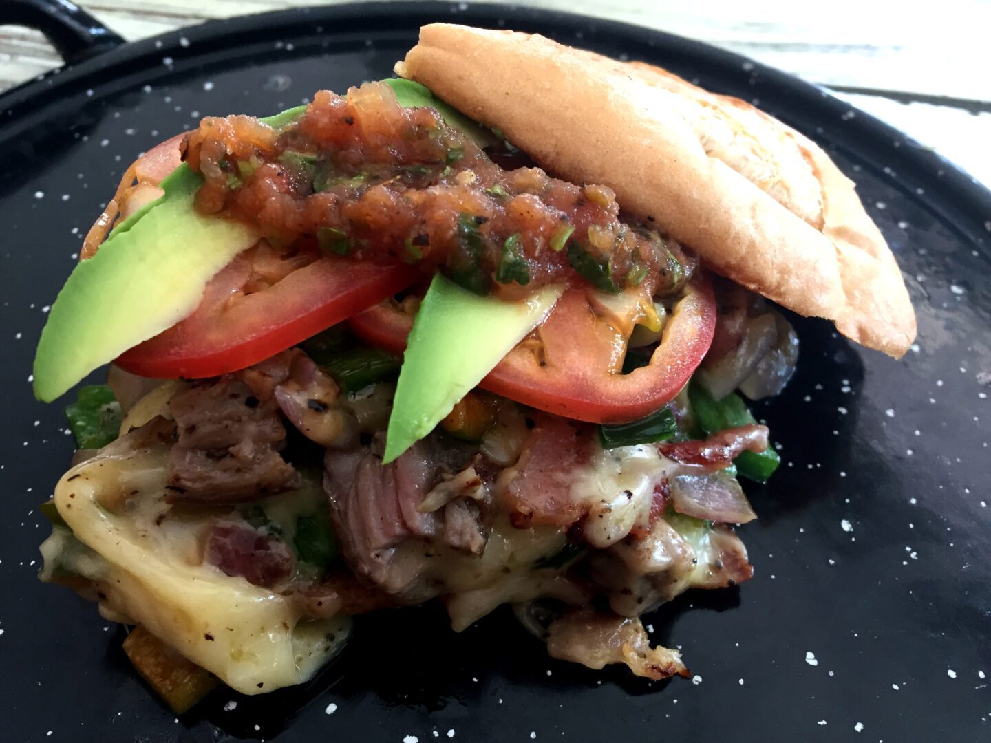 A sandwich with meat, tomatoes and avocado on a black plate.
