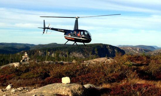 A helicopter flies over a rocky area with mountains in the background.